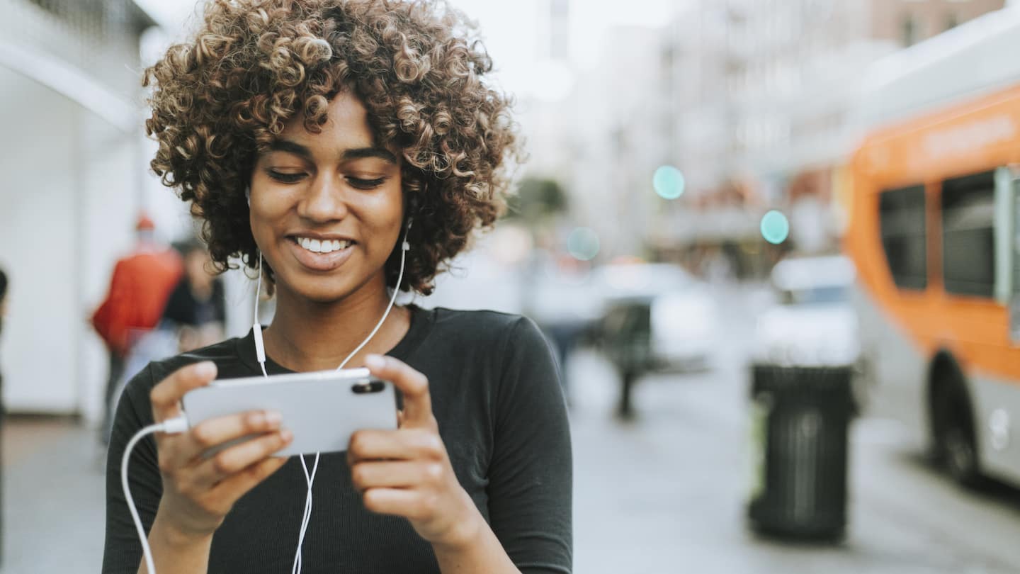 Image of smiling woman wearing headphones and interacting with her phone on a busy city street.