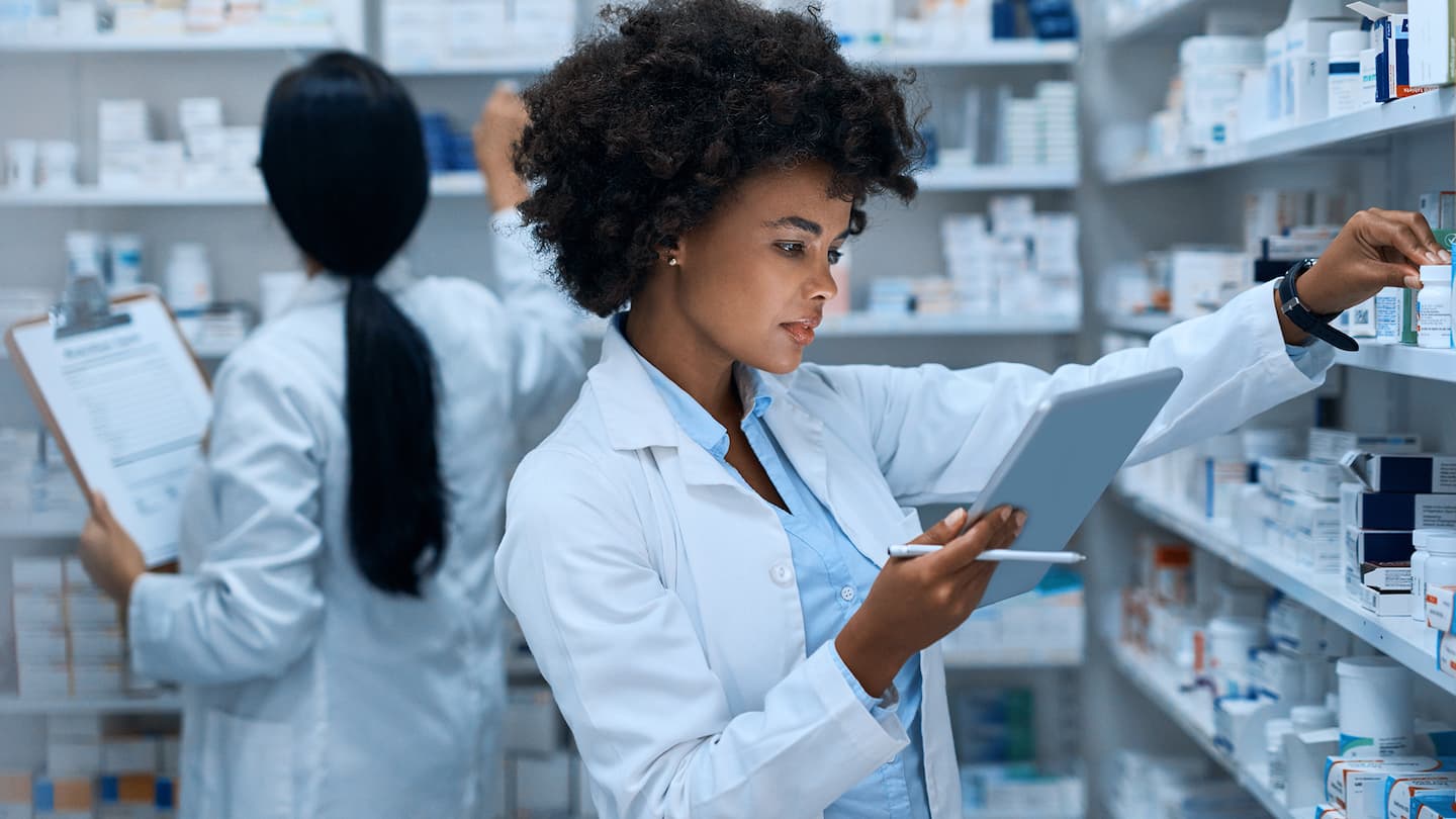 Imagem de farmacêuticos usando jalecos de laboratório brancos verificando o inventário em uma sala cheia de prateleiras de medicamentos.